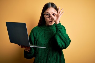 Poster - Young asian business woman wearing glasses and working using computer laptop doing ok gesture shocked with surprised face, eye looking through fingers. Unbelieving expression.