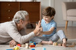 © fizkes - Overjoyed senior grandfather lying on floor in living room play with blocks with cute little grandson, happy mature grandparent relax engaged in game with building bricks with small grandchild at home