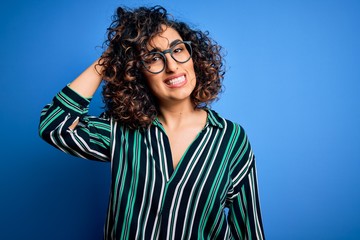 Poster - Young beautiful curly arab woman wearing striped shirt and glasses over blue background confuse and wonder about question. Uncertain with doubt, thinking with hand on head. Pensive concept.