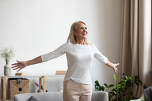 Overjoyed Mature Grandmother Standing With Outstretched Arms Near Comfortable Couch, Breathing Fresh Air, Enjoying Freedom, Happy Life Moment. Smiling Older Woman Feeling Thankful For Good Day.