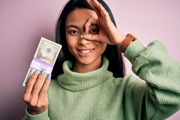 Wall Mural - Young beautiful chinese woman holding dollars standing over isolated pink background with happy face smiling doing ok sign with hand on eye looking through fingers