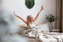 Overjoyed Pleasant Middle Aged Woman Sitting In Bed After Waking Up, Stretching Muscles After Good Night Rest. Joyful Healthy Older Female Retiree Greeting New Day, Feeling Energetic In Morning.