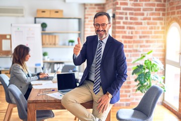 Middle age handsome businessman wearing glasses sitting on desk at the office doing happy thumbs up gesture with hand. Approving expression looking at the camera with showing success.