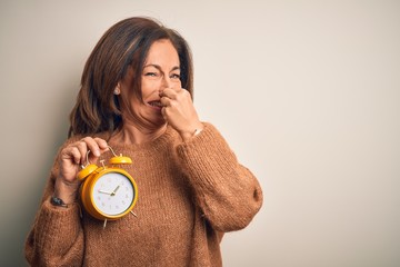 Poster - Middle age brunette woman holding clasic alarm clock over isolated background smelling something stinky and disgusting, intolerable smell, holding breath with fingers on nose. Bad smell