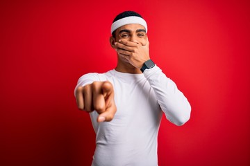 Young handsome african american sportsman wearing sportswear over red background laughing at you, pointing finger to the camera with hand over mouth, shame expression