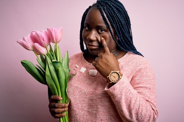 Wall Mural - Young african american plus size woman with braids holding bouquet of pink tulips flower Pointing to the eye watching you gesture, suspicious expression