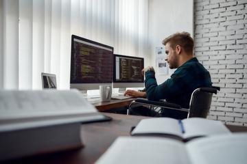 Computer programmer at work. Side view of young web developer in a wheelchair writing code on multiple computer screens while sitting at his workplace in the modern office. IT software