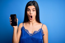 Young Beautiful Brunette Woman Wearing Blue Lingerie Holding Smartphone Showing Screen Scared In Shock With A Surprise Face, Afraid And Excited With Fear Expression