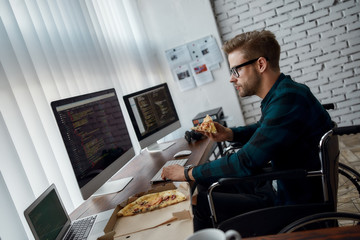 Focused male web developer in a wheelchair eating pizza and writing program code while sitting at workplace with three monitors in the modern office. Lunch time