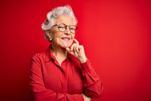 Senior Beautiful Grey-haired Woman Wearing Casual Shirt And Glasses Over Red Background Looking Confident At The Camera With Smile With Crossed Arms And Hand Raised On Chin. Thinking Positive.