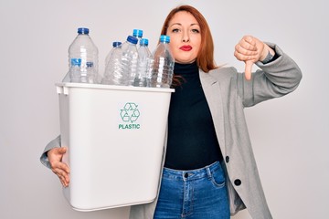 Poster - Young beautiful redhead woman recycling holding trash can with plastic bottles to recycle with angry face, negative sign showing dislike with thumbs down, rejection concept