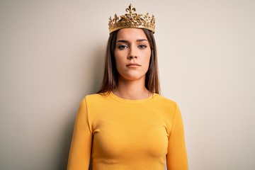 Wall Mural - Young beautiful brunette woman wearing golden queen crown over white background with serious expression on face. Simple and natural looking at the camera.