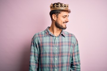 Wall Mural - Young man with beard wearing golden crown of king standing over isolated pink background looking away to side with smile on face, natural expression. Laughing confident.