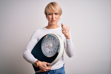 Wall Mural - Young blonde woman with short hair holding scale for healthy weight and lifestyle annoyed and frustrated shouting with anger, crazy and yelling with raised hand, anger concept