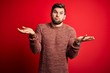 © Krakenimages.com - Young blond man with beard and blue eyes wearing casual sweater over red background clueless and confused expression with arms and hands raised. Doubt concept.