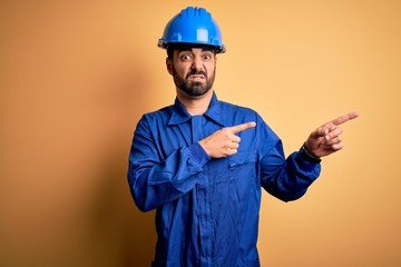 Wall Mural - Mechanic man with beard wearing blue uniform and safety helmet over yellow background Pointing aside worried and nervous with both hands, concerned and surprised expression