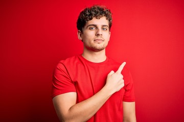 Wall Mural - Young blond handsome man with curly hair wearing casual t-shirt over red background Pointing with hand finger to the side showing advertisement, serious and calm face