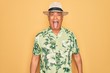 Middle age senior grey-haired man wearing summer hat and floral shirt on beach vacation sticking tongue out happy with funny expression. Emotion concept.