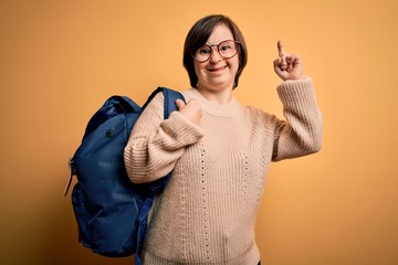 Sticker - Young down syndrome student woman wearing school bag over yellow background surprised with an idea or question pointing finger with happy face, number one