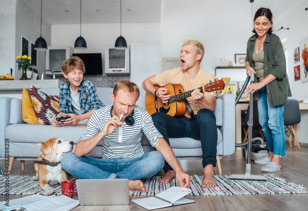 Quarantine time: Family at living room together. Father distance working using laptop, mother cleaning the room, older son playing acoustic guitar and singing a songs, little son playing video console - obrazy, fototapety, plakaty 
