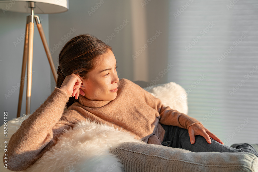 Staying home Asian woman relaxing pensive looking out the window sitting in living room sofa enjoying free time indoors during coronavirus outbreak. - obrazy, fototapety, plakaty 
