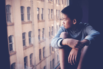 Wall Mural - Sad Asian preteen boy feeling lonely, looking out of window from his room, social distancing, isolation, mental health concept