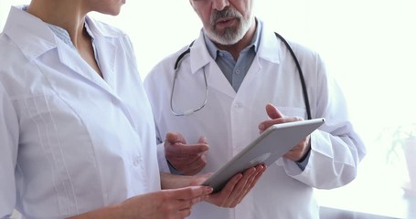 Wall Mural - Medical team two old male and young female doctors wear white coats talk, use digital tablet, handshake. 2 hospital workers shake hands. Partnership in medicine concept, healthcare tech. Close up view