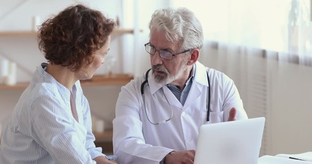 Wall Mural - Old male doctor consulting female patient showing medical test results on laptop. Senior 60s physician explaining treatment talking to 30s woman client during checkup visit. Women healthcare concept.