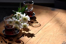 Spotlight On The Wooden Table With Tea In Tall Glass Cups And White Daisy Flowers In Back Light. Copy Space.