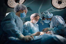 Medical Team In The Operating Room, Dark Background. The Theater Of The Operating Room, An International Team Of Professional Doctors In A Modern Operating Room Are Conducting An Operation. Saving