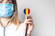 female doctor in a medical mask holds a stethoscope on a light background. Added flag of Romania. Concept medicine, level of medicine, virus, epidemic