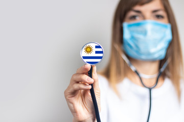 female doctor in a medical mask holds a stethoscope on a light background. Added flag of Uruguay. Concept medicine, level of medicine, virus, epidemic