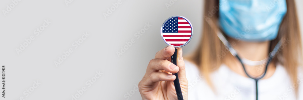 female doctor in a medical mask holds a stethoscope on a light background. Added flag of United States of America. Concept medicine, level of medicine, virus, epidemic. Baner - obrazy, fototapety, plakaty 