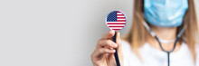 Female Doctor In A Medical Mask Holds A Stethoscope On A Light Background. Added Flag Of United States Of America. Concept Medicine, Level Of Medicine, Virus, Epidemic. Baner