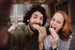 © StratfordProductions - Cheerful young professional couple taking a selfie using smartphone while grabbing a bite of sandwich