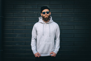 Wall Mural - City portrait of handsome hipster guy with beard wearing gray blank hoodie or hoody and hat with space for your logo or design. Mockup for print