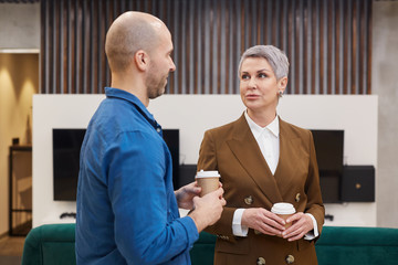 Wall Mural - Waist up portrait of modern mature businesswoman talking to colleague and holding coffee cup during break in office lobby