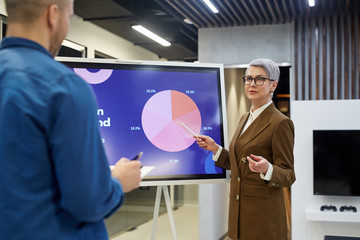 Wall Mural - Portrait of modern mature businesswoman pointing at pie chart while giving presentation for employees or clients in office hall, copy space