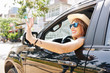 Beautyiful smiling young woman in hat and sunglasses looking out of car window and waving with hand
