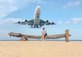 Woman sitting relax on old wood log tree enjoy on the beach while passenger airplane landing above in blue sky at Mai Khao beach Phuket Thailand travel destinations concept.