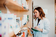 .Young female pharmacist working in her large pharmacy. Placing medications, taking inventory with her green notebook. Lifestyle