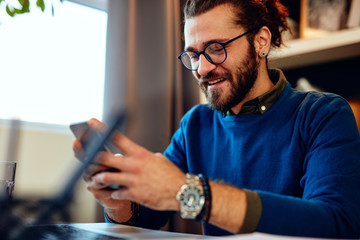 Attractive caucasian bearded entrepreneur sitting in his office, using smart phone and taking a break from work.	