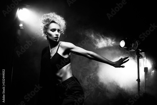 Cuadros en Lienzo A beautiful blonde girl with an elegant hairstyle and large breasts, wearing a bra, trousers and a blazer, artistically poses in the rays of spotlights in the smoke