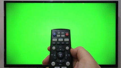 Person pointing a TV remote and pressing buttons in front of a TV with green screen