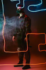 armed mixed race cyberpunk player in protective mask standing near neon lighting on black with smoke