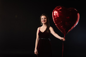 Wall Mural - smiling woman in dress holding heart-shaped balloon isolated on black