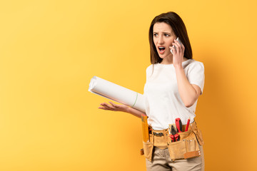 Wall Mural - shocked handywoman talking on smartphone and holding project on yellow background