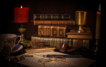 An Old Desk With A Pile Of Vintage Brown Leather Books, An Open Book With Eye Glasses, A Cup Of Coffee, A Red Candle With Flame, A Vintage Pocket Clock, Earth Globe.