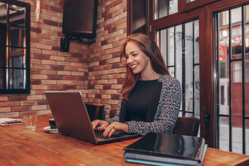 Happy young business woman using computer and smiling while sitting in cafe.