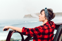 Side View Of Extraordinary Woman With Black Short Hair And Bandana On Head And Piercing In Red Plaid Shirt Leaning On Car And Looking Away At Seaside